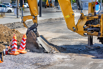 The bucket of a large construction excavator lifts a layer of old asphalt on the road against the backdrop of a city street on a sunny summer day. - 634710644