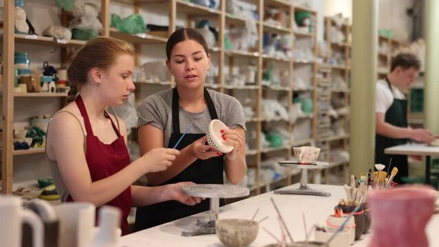 Young woman teacher showing teenage boy and girl how to paint ceramic cup