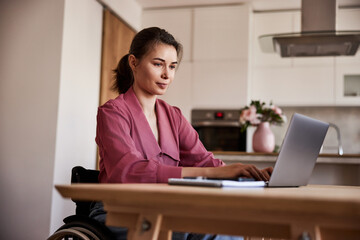 A pretty woman in a wheelchair working over a laptop at the wooden table at home.