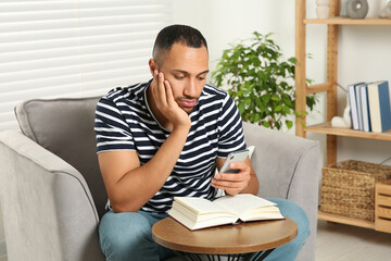 Young man using smartphone while reading book at home. Internet addiction
