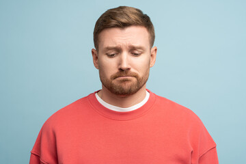Fototapeta premium Frustrated unhappy man sadly looks down with upset expression isolated on studio wall. Concept of depression, disappointment. Regretful tired male with difficult trouble slack of money, broken heart