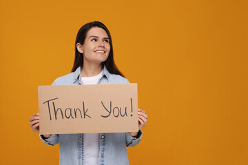 Happy woman holding cardboard sheet with phrase Thank You on orange background, space for text
