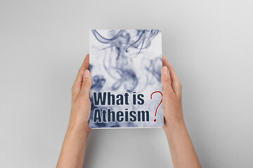 Woman holding book about atheism on light background, top view