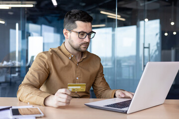 A serious young male businessman works in the office at a laptop and conducts transactions with a credit card in his hand