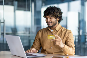 A young businessman inside the office at the workplace uses a laptop and a bank credit card, the man makes purchases in an online store and transfers money