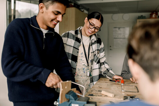 Happy female teacher standing with male student cutting wood during carpentry class