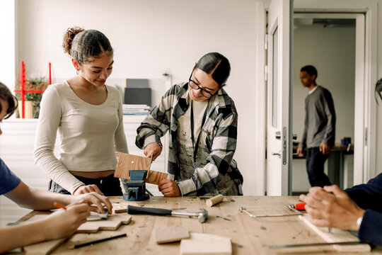 Teacher helping female teenage student while cutting wood during carpentry class