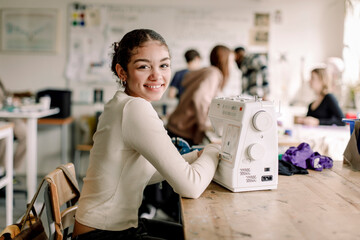 Side view portrait of smiling teenage girl with sewing machine on desk at high school