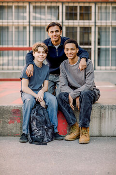 Portrait of smiling teenage student with arms around male friends sitting on concrete in high school campus