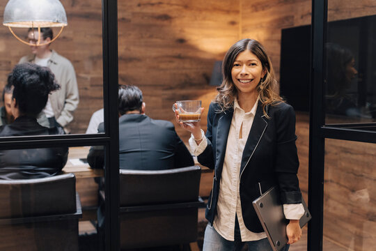Portrait of smiling businesswoman holding coffee cup and laptop standing at doorway with colleagues in board room