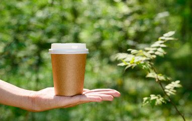 Paper disposable cup on the hand against the background of green foliage. The concept of using eco-friendly tableware made from biodegradable materials. Template for logo or advertising