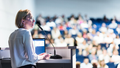 Female speaker giving a talk on corporate business conference. Unrecognizable people in audience at...