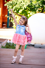 Cute little girl holding a pink backpack, playing in the yard. Beautiful little girl smiles in a smart dress. Preschool education, preparation for school.