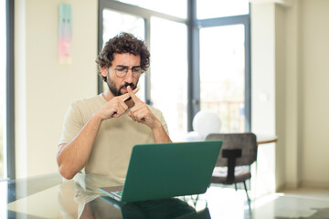 young adult bearded man with a laptop looking serious and displeased with both fingers crossed up...