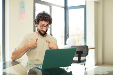 young adult bearded man with a laptop feeling happy, surprised and proud, pointing to self with an excited, amazed look