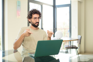 young adult bearded man with a laptop pointing forward at camera with both fingers and angry expression, telling you to do your duty