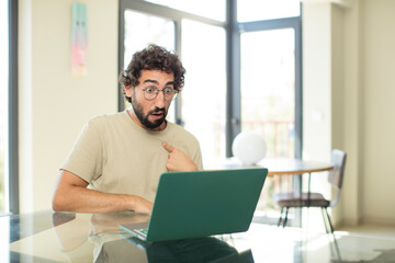 young adult bearded man with a laptop looking shocked and surprised with mouth wide open, pointing to self