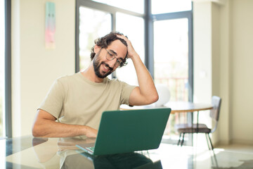 young adult bearded man with a laptop smiling cheerfully and casually, taking hand to head with a positive, happy and confident look