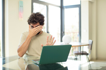 young adult bearded man with a laptop covering face with hand and putting other hand up front to...