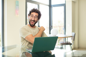 young adult bearded man with a laptop feeling happy, satisfied and powerful, flexing fit and muscular biceps, looking strong after the gym