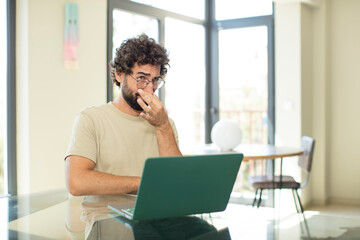 young adult bearded man with a laptop feeling disgusted, holding nose to avoid smelling a foul and unpleasant stench