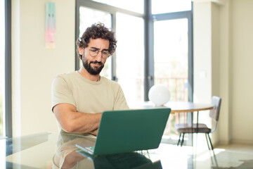 young adult bearded man with a laptop feeling displeased and disappointed, looking serious, annoyed and angry with crossed arms