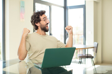 young adult bearded man with a laptop feeling happy, positive and successful, celebrating victory, achievements or good luck