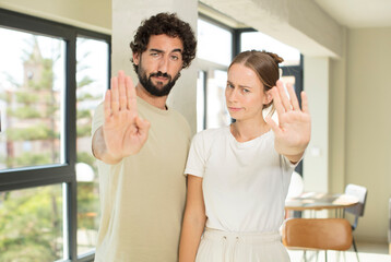 young adult couple looking serious, stern, displeased and angry showing open palm making stop...