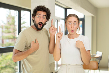 young adult couple feeling proud and surprised, pointing to self confidently, feeling like successful number one