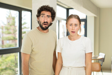 young adult couple feeling terrified and shocked, with mouth wide open in surprise