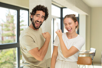 young adult couple feeling happy, satisfied and powerful, flexing fit and muscular biceps, looking...
