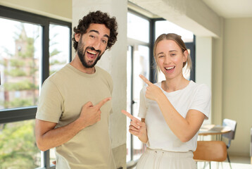 young adult couple smiling cheerfully and pointing to copy space on palm on the side, showing or advertising an object