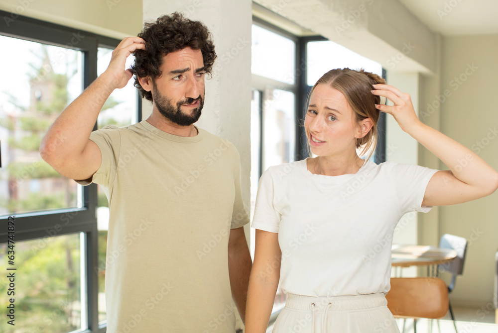 Wall mural young adult couple feeling puzzled and confused, scratching head and looking to the side