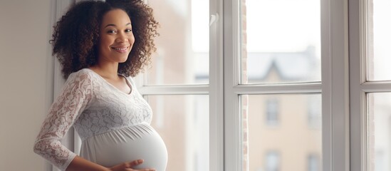 Young pregnant black woman smiling and looking at empty space enjoying her pregnancy near a window at home - obrazy, fototapety, plakaty