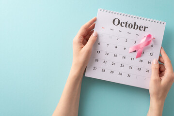 Preparing for my check-up: First person top view of my hands holding an October calendar, planning the doctor's visit for breast cancer awareness. Pink ribbon on pastel blue backdrop with empty space