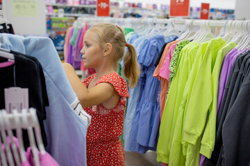 A little blonde girl chooses a new dress in a clothing store. Examines things hanging on a hanger. The concept of independent wardrobe choice by children