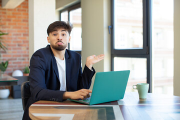 young handsome man shrugging, feeling confused and uncertain. freelance concept with laptop