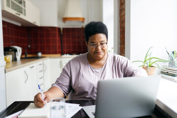 African plus size aged woman sitting in front of laptop planning purchases, making list before going to supermarket, using online application to compare prices, searching for sales, special offers - obrazy, fototapety, plakaty