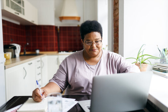 African plus size aged woman sitting in front of laptop planning purchases, making list before going to supermarket, using online application to compare prices, searching for sales, special offers