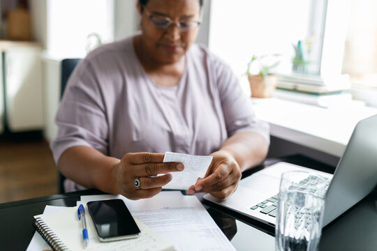 Selective focus on black female hands holding utility bill or paycheck sitting in front of laptop with smartphone on table near window at kitchen, puzzled with high prices due to inflation