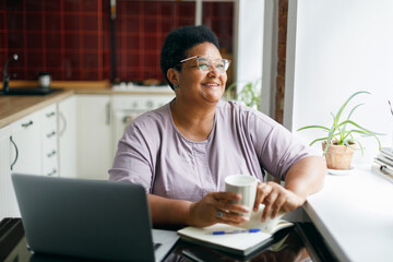 Cheerful happy smiling overweight mature black woman in spectacles sitting at kitchen table with...