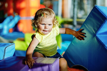 Obraz na płótnie Canvas Happy blond little toddler girl having fun and sliding on indoor playground at daycare or nursery. Positive funny baby child smiling. Healthy girl climbing on slide.