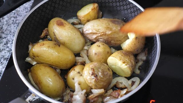 Cooking homemade meals. Close-up of appetizing jacket potatoes in process of frying in pan in oil with bacon slices and onions being mixed with wooden spatula. High quality 4k footage