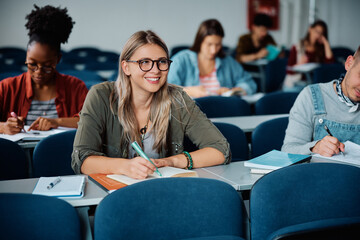Happy female student taking notes while attending lecture at university. - obrazy, fototapety, plakaty