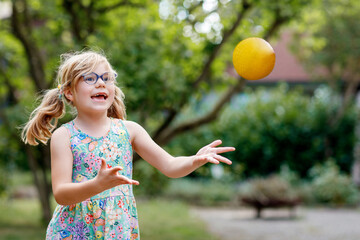 Little preschool girl with eyeglasses playing with ball outdoors. Happy smiling child catching and throwing, laughing and making sports. Active leisure with children and kids. Summer day on backyard