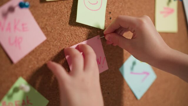 Little child pins memo stick with words Pop Corn to cork-board in kitchen closeup. Small girl leaves reminder about favorite snack on board at home