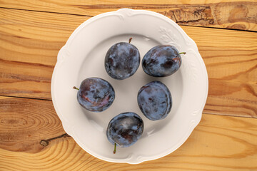 Several sweet ripe plums with white ceramic plate on wooden table, macro, top view.