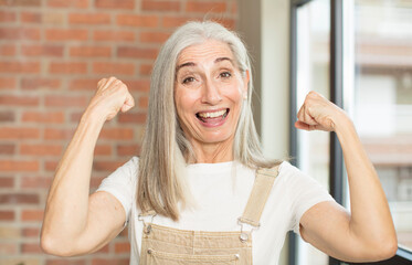 senior pretty woman feeling happy, satisfied and powerful, flexing fit and muscular biceps, looking...