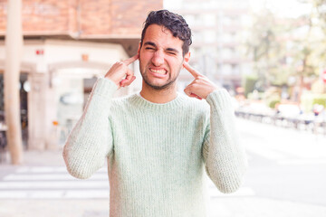 young hispanic man looking angry, stressed and annoyed, covering both ears to a deafening noise,...
