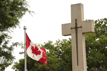 canadian flag blowing in wind next to metal cross fastened to stone cross monument with trees and...
